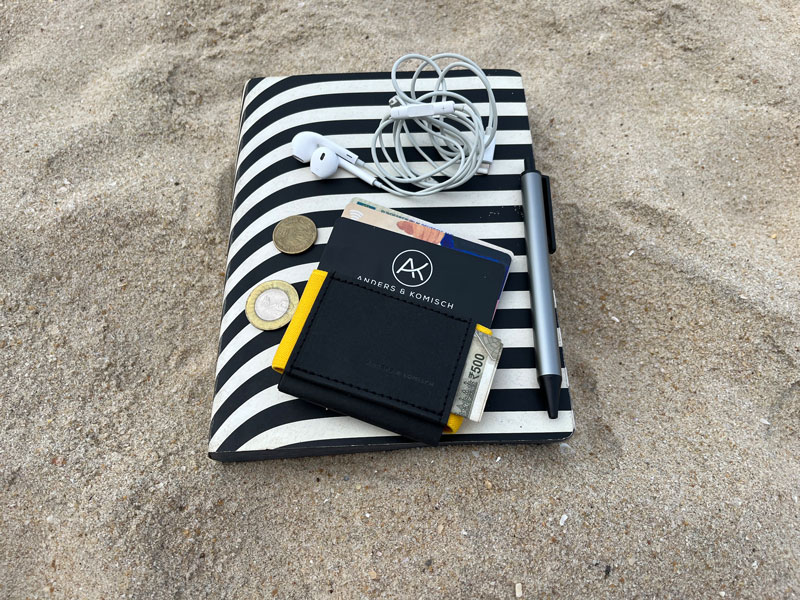 Das A&K Mini travel wallet für die nächste Reise liegt auf einem Journal im Sand. Es enthält Scheine, Karten und Münzen. Als Größenvergleich liegen Kopfhörer daneben