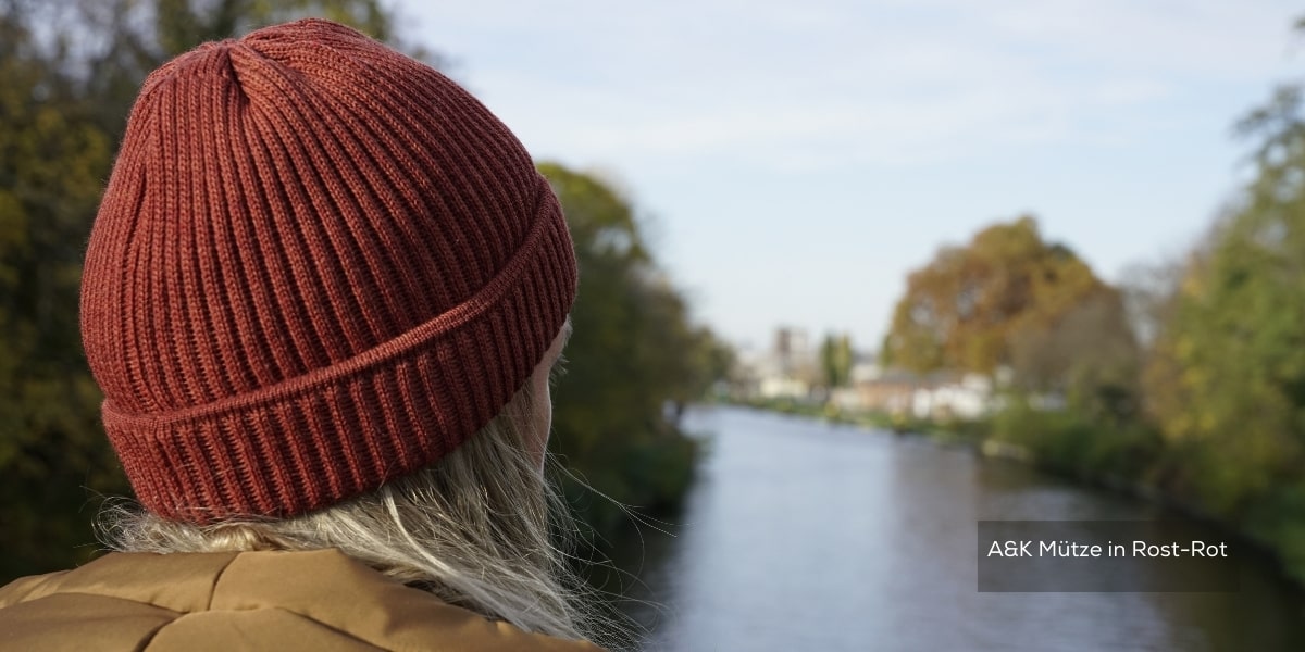 Frau mit langen Haaren trägt eine Rost-Rote Mütze und blickt auf einen Fluss.